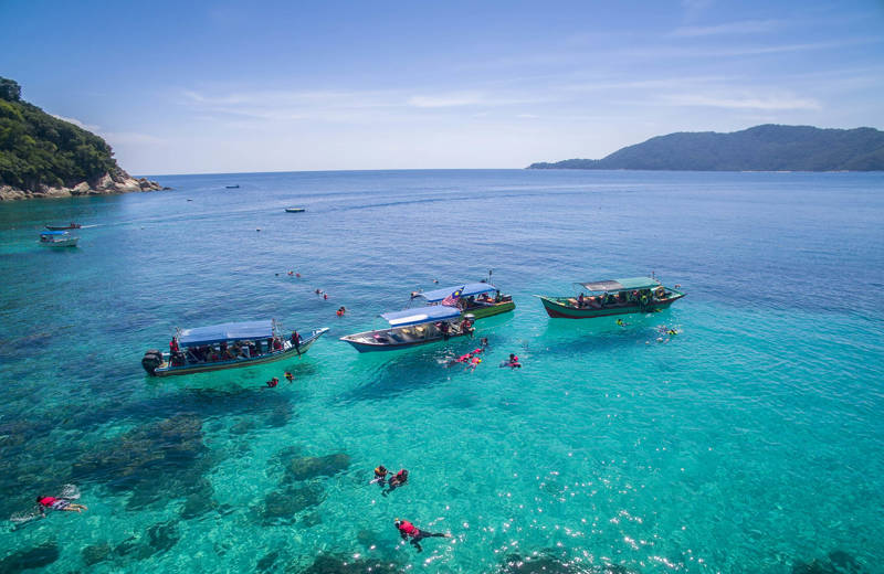 Een paar boten voor de kust van de Perhentian-eilanden, met mensen die duiken en snorkelen in het lichtblauwe water rondom de boten.