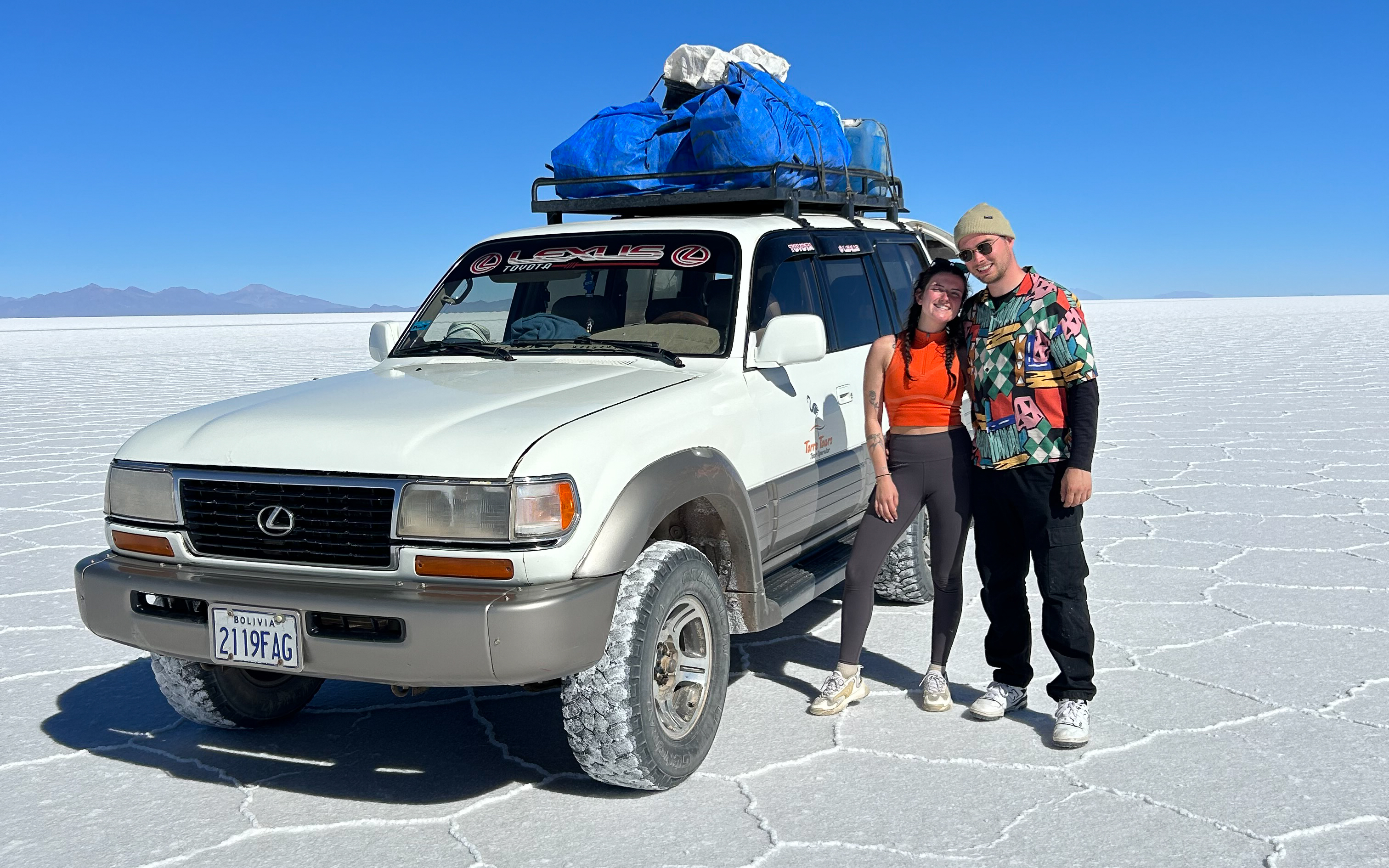 Een jong koppel poseert bij een witte 4x4 terreinwagen, volgeladen met bagage op het dak, midden op de uitgestrekte, witte zoutvlaktes van Salar de Uyuni in Bolivia. - KILROY
