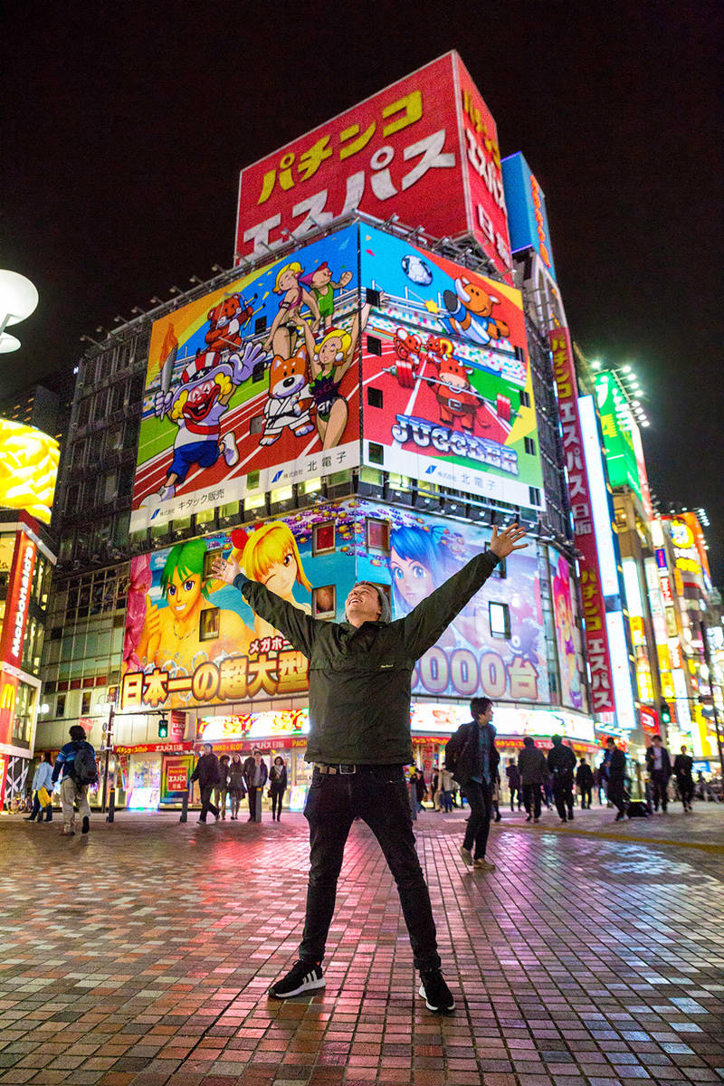 Man spreidt zijn armen op een plein in Tokio met grote billboards met Japanse animatiefiguren op gebouwen op de achtergrond