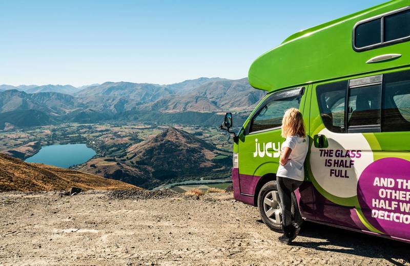 Een meisje leunt tegen een Jucy Condo camper, ze kijkt naar een meer en bergen.