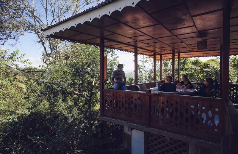 Mensen zitten een rond tafel op een overdekt terras, omgeven door groene natuur