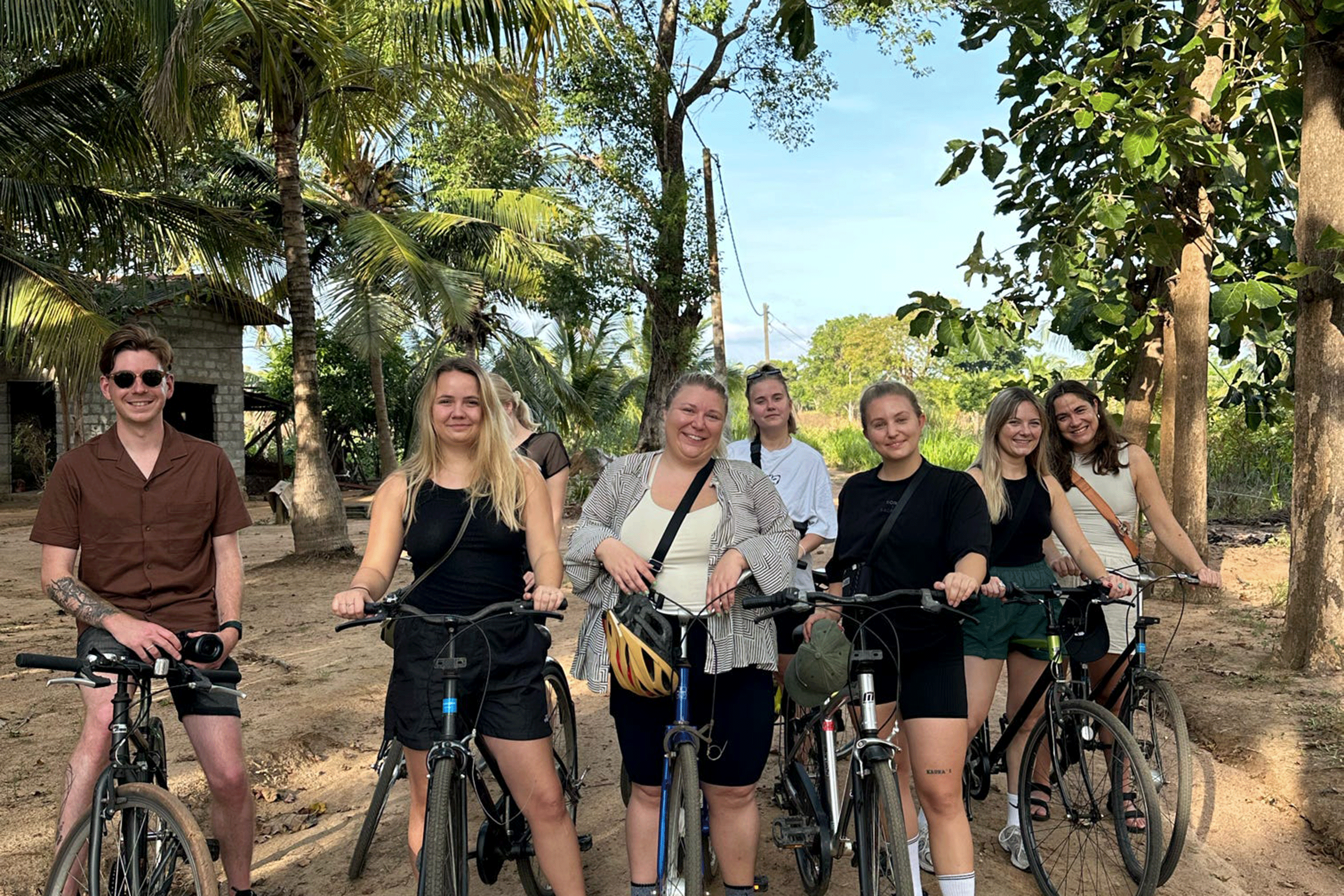 Een groep lachende backpackers met hun fietsen tijdens een excursie in een tropische, groene omgeving met palmbomen.