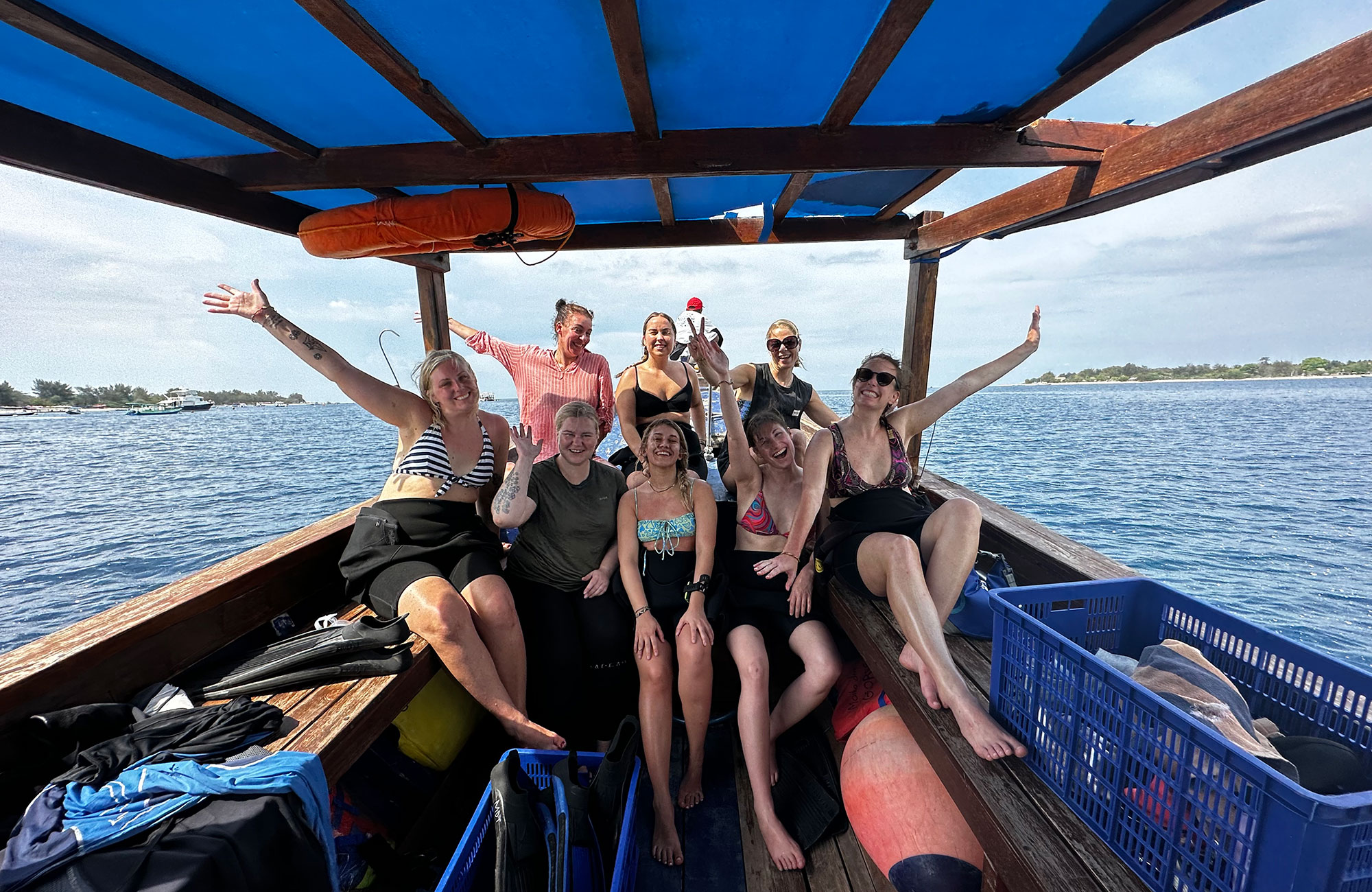 Groepsfoto van een diverse groep lachende en zwaaiende mensen op een houten boot met een blauwe kap, op de turquoise oceaan bij een tropisch eiland in Bali.