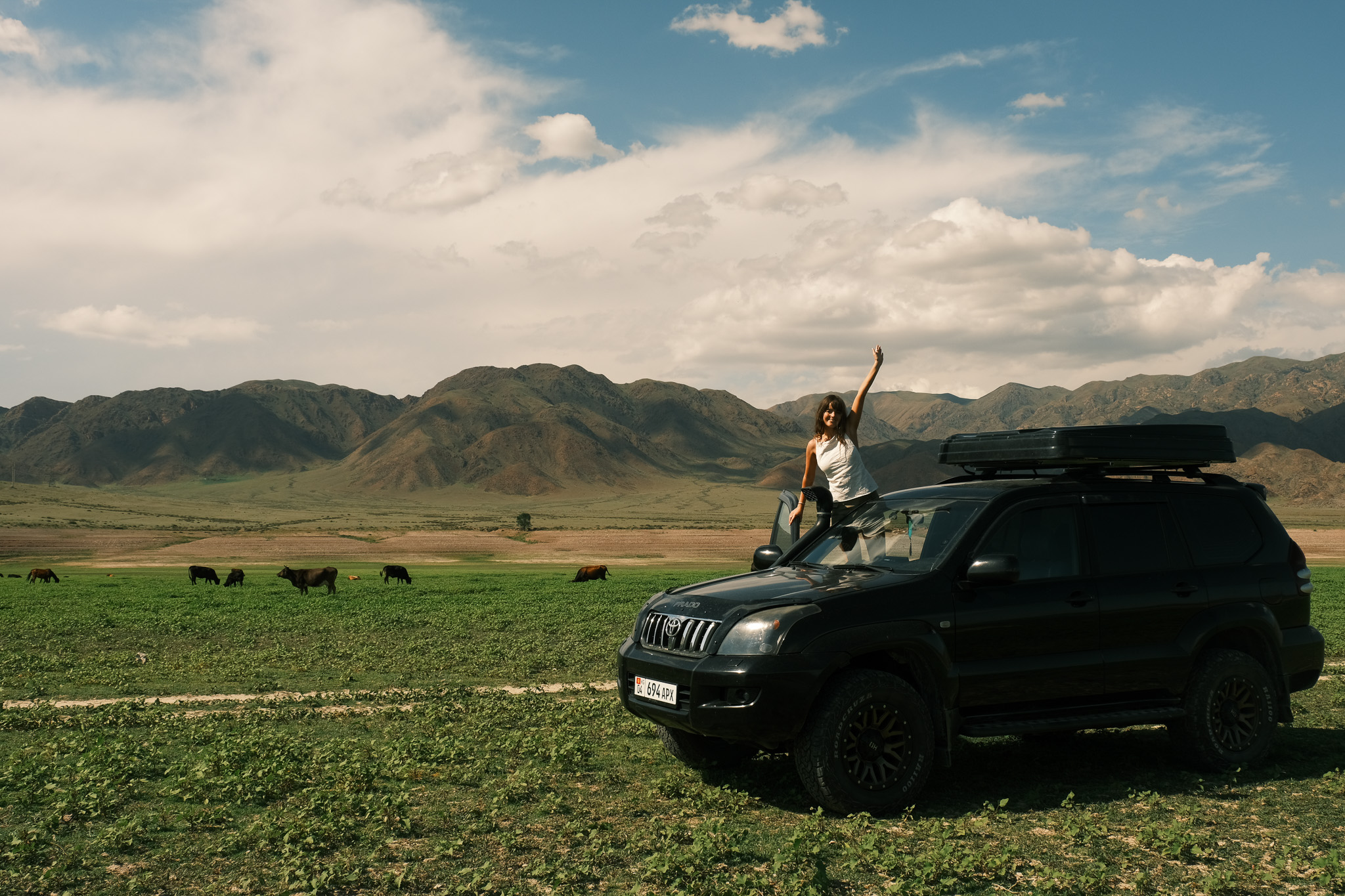Een reiziger zwaait vanuit het open dak van een 4x4 met daktent in het landschap van Kirgizië.