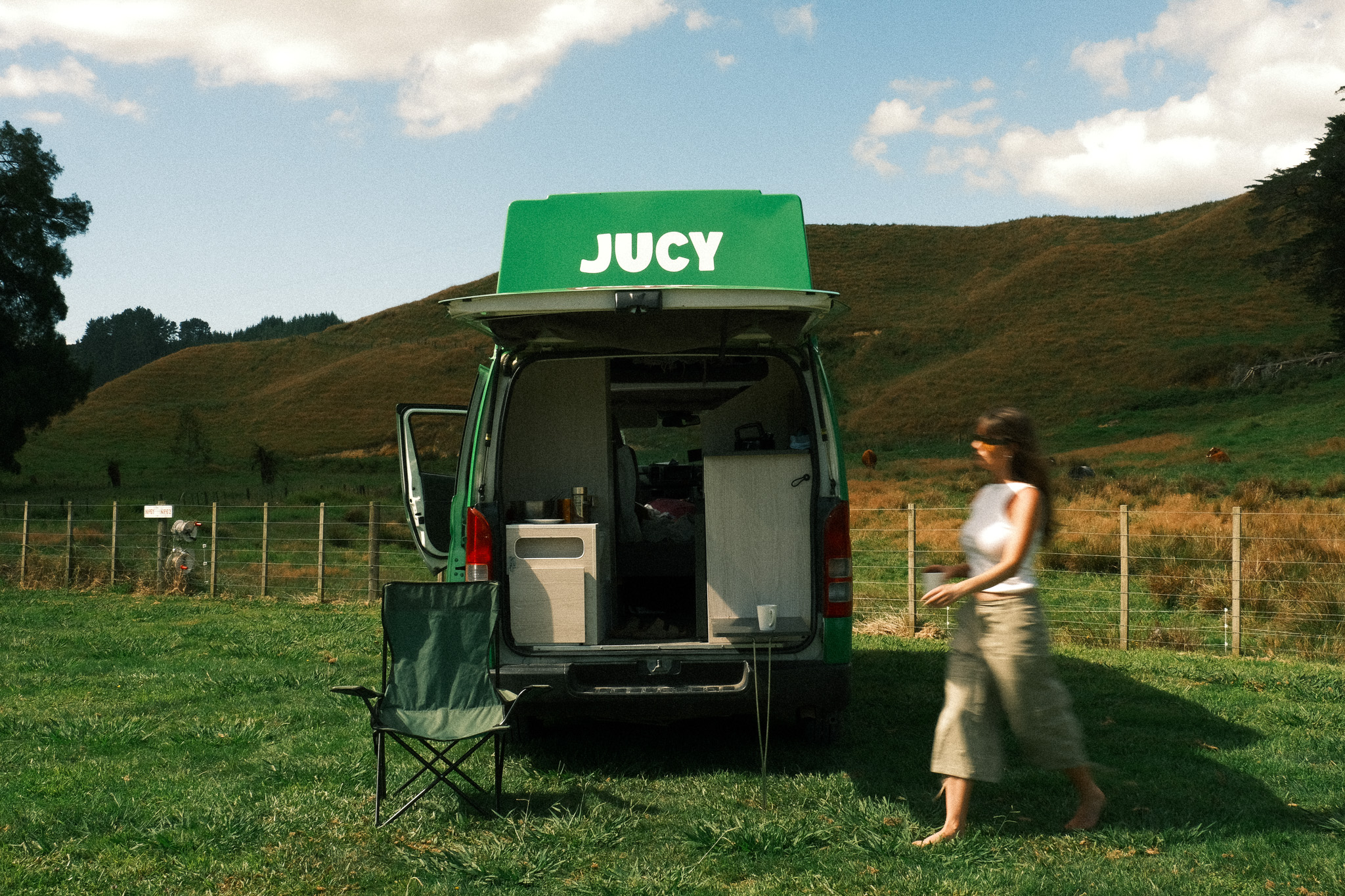 JUCY-campervan geparkeerd op een grasveld in het Nieuw-Zeelandse platteland, met open achterdeuren, een kampeerstoel en glooiende groene heuvels op de achtergrond.