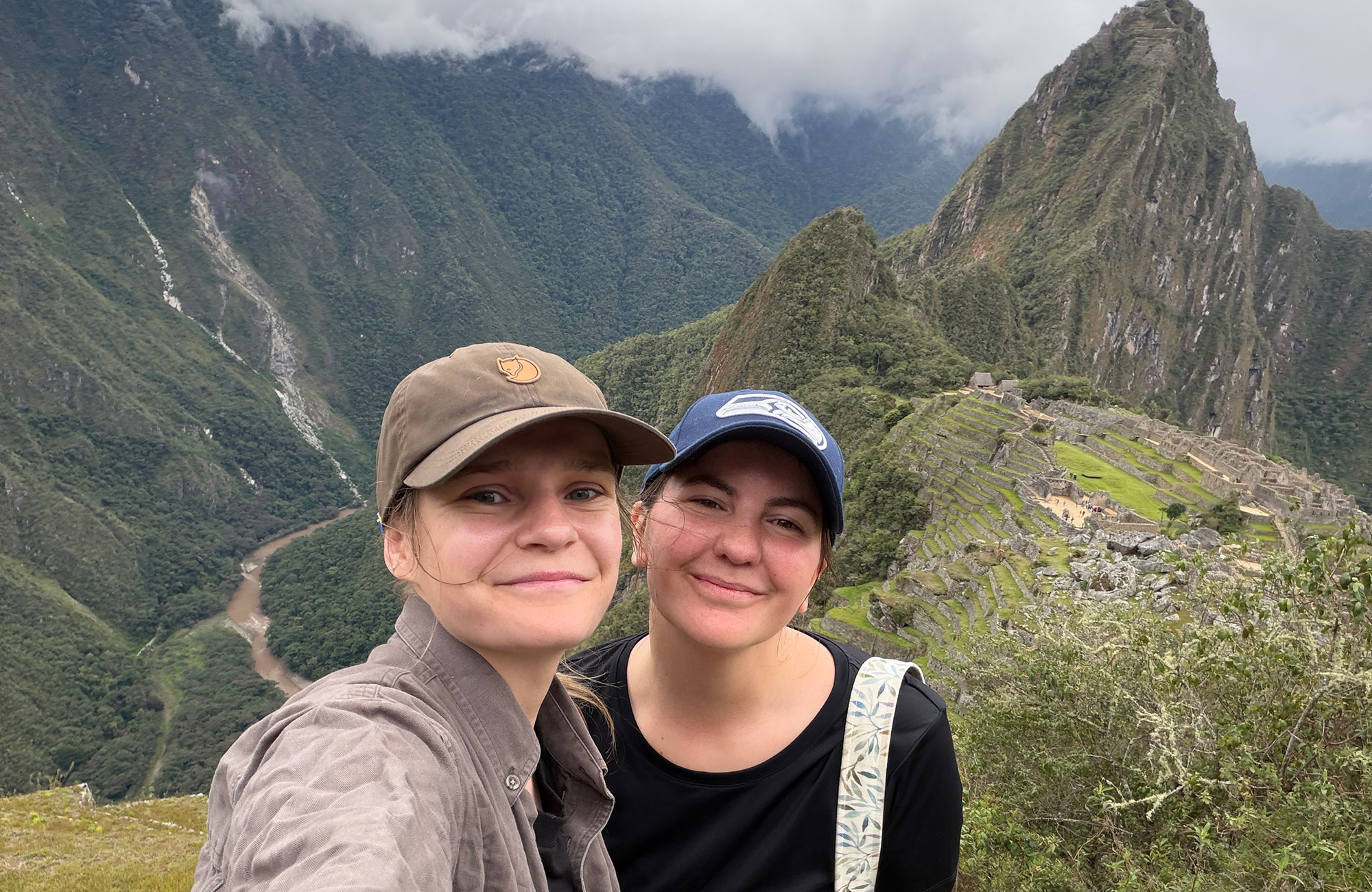 Selfie van twee glimlachende vrouwen met petjes op, met op de achtergrond de historische ruïnes en groene bergen van Machu Picchu in de mist.