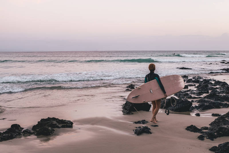 Iemand loopt op het strand met een surfboard onder zijn arm richting de zee
