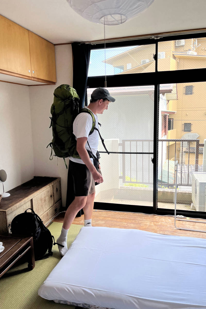 Man met grote rugzak staat in een slaapkamer in zijn Japanse accommodatie en kijkt rond