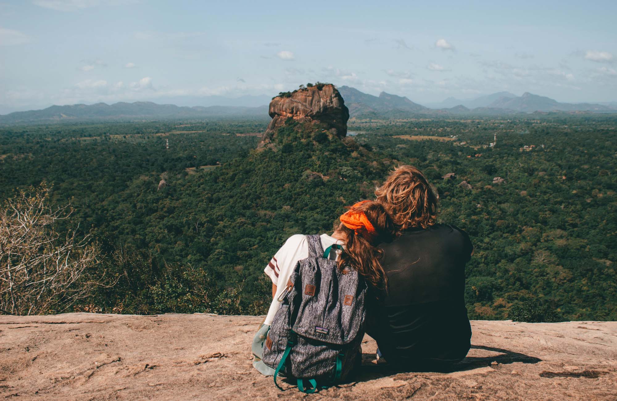 Koppel, gezien vanaf de rug, rustend op een rotsplateau en uitkijkend over de dichte jungle en de iconische Sigiriya Rock Fortress in Sri Lanka. - KILROY
