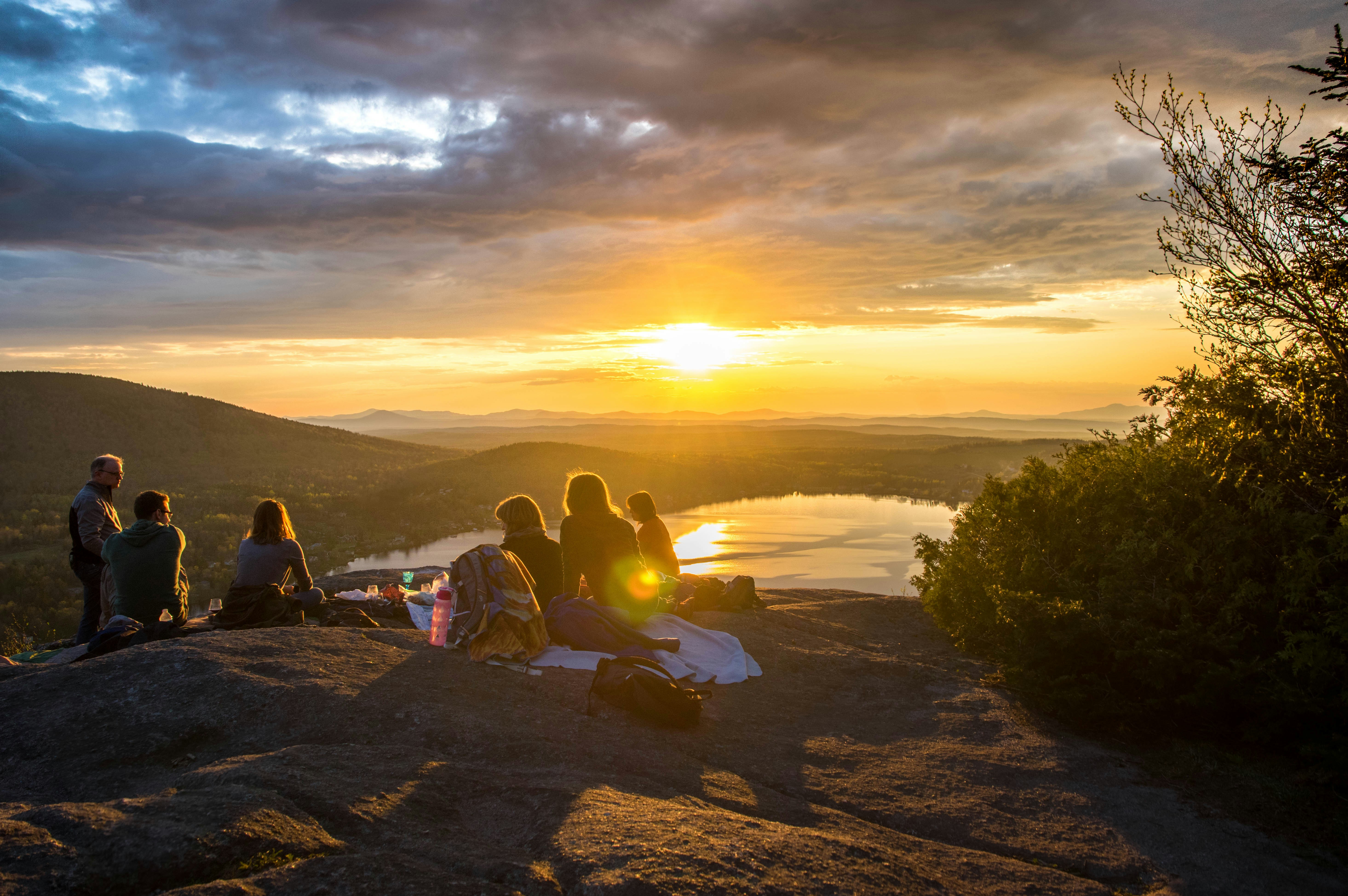 Een groep jonge reizigers zit op een rotswand en geniet van een picknick bij zonsondergang met uitzicht over een meer en beboste heuvels.