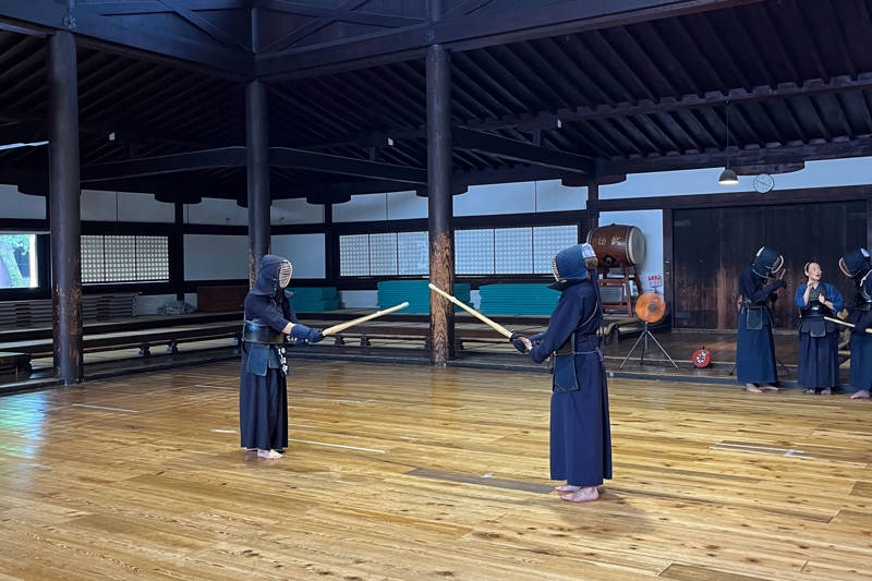 Twee mensen in samoeraikleding staan in een typische Japanse tempel met zwaarden in hun handen op een houten vloer.